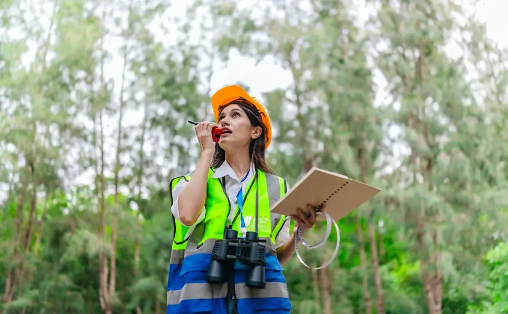 Female environmental surveyor observes forest conditions using binoculars, takes notes, communicates data. 