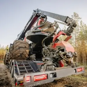 Heavy logging machinery on a trailer, set against a forest backdrop with pine trees under a clear sky, ready for timber harvesting.